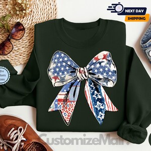 May include: A dark green sweatshirt with a large red, white, and blue bow with stars and stripes. The bow is in the center of the sweatshirt. The sweatshirt is on a white surface.