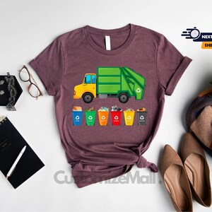 May include: A heather maroon t-shirt with a graphic of a green garbage truck and colorful recycling bins. The text "Next Day Shipping" is visible in the top right corner of the image.