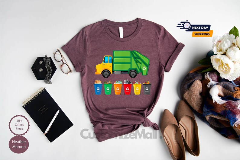May include: A heather maroon t-shirt with a graphic of a green garbage truck and colorful recycling bins. The text "Next Day Shipping" is visible in the top right corner of the image.