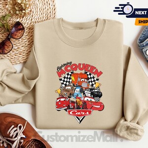 a beige sweatshirt with a graphic of Lightning McQueen and various cars from the Cars movie, along with sunglasses and a pair of jeans.