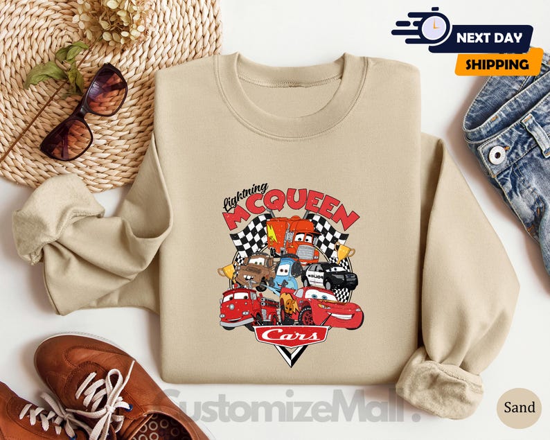 a beige sweatshirt with a graphic of Lightning McQueen and various cars from the Cars movie, along with sunglasses and a pair of jeans.