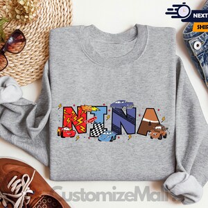 Puede incluir: Una sudadera gris con el nombre "NINA" escrito en letras de colores con coches de dibujos animados y temas de carreras. La sudadera es de color gris deportivo.
