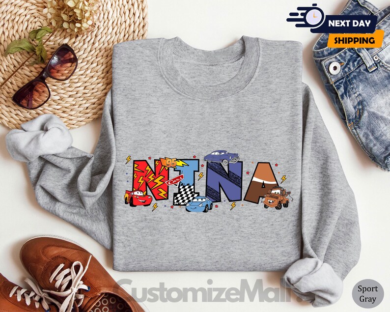 Puede incluir: Una sudadera gris con el nombre "NINA" escrito en letras de colores con coches de dibujos animados y temas de carreras. La sudadera es de color gris deportivo.