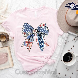 May include: A pink t-shirt with a large, colorful bow design. The bow is made up of red, white, and blue fabric with a star pattern. The shirt is a light pink color and has a crew neck.