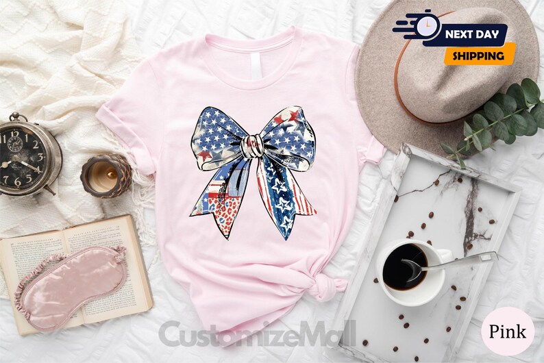 May include: A pink t-shirt with a large, colorful bow design. The bow is made up of red, white, and blue fabric with a star pattern. The shirt is a light pink color and has a crew neck.
