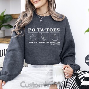 May include: Dark heather gray sweatshirt with the text "PO*TA*TOES" and illustrations of cooking potatoes. The text below reads "BOIL 'EM MASH 'EM STICK 'EM IN A STEW". The sweatshirt is a comfortable, casual style.