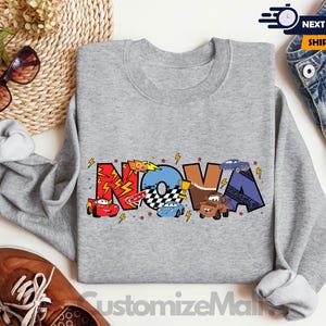 Może przedstawiać: Szara bluza z napisem "NOVA" w kolorowych literach z kresk&oacute;wkowymi samochodami i błyskawicami. Bluza ma okrągły dekolt.