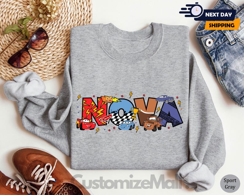 Może przedstawiać: Szara bluza z napisem "NOVA" w kolorowych literach z kresk&oacute;wkowymi samochodami i błyskawicami. Bluza ma okrągły dekolt.