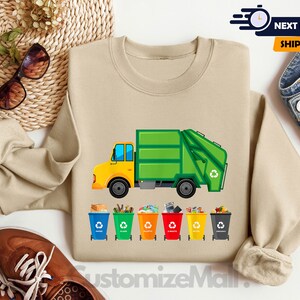 May include: A tan sweatshirt with a green and yellow garbage truck and five colorful recycling bins with the words "Paper", "Plastic", "Glass", "Metal", and "Organic" printed on them.