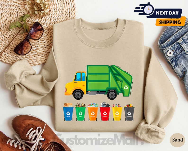 May include: A tan sweatshirt with a green and yellow garbage truck and five colorful recycling bins with the words "Paper", "Plastic", "Glass", "Metal", and "Organic" printed on them.