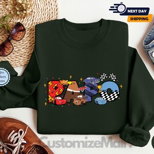 Puede incluir: Una sudadera de color verde oscuro con un gr&aacute;fico de los personajes de Cars, incluyendo Rayo McQueen, Mate y otros. El gr&aacute;fico est&aacute; en un estilo de dibujos animados colorido e incluye la palabra "RACO" en una fuente estilizada.
