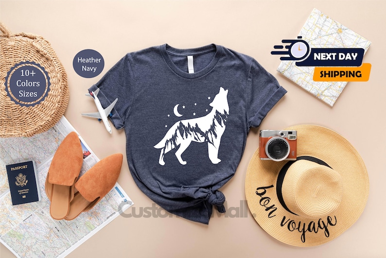 K&ouml;nnte beinhalten: Dunkelblau meliertes T-Shirt mit einem wei&szlig;en Wolfsdesign, Mond und Sternen. Das Bild zeigt Reiseaccessoires, eine Karte, einen Reisepass und einen Strohhut mit der Aufschrift "bon voyage".