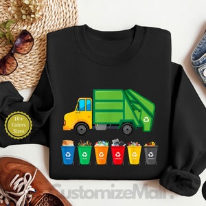 May include: Black sweatshirt with a green garbage truck and five colorful recycling bins. The truck has a yellow cab and the text "10+ Colors Sizes" is on the left side of the image.