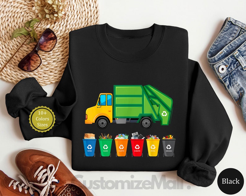 May include: Black sweatshirt with a green garbage truck and five colorful recycling bins. The truck has a yellow cab and the text "10+ Colors Sizes" is on the left side of the image.