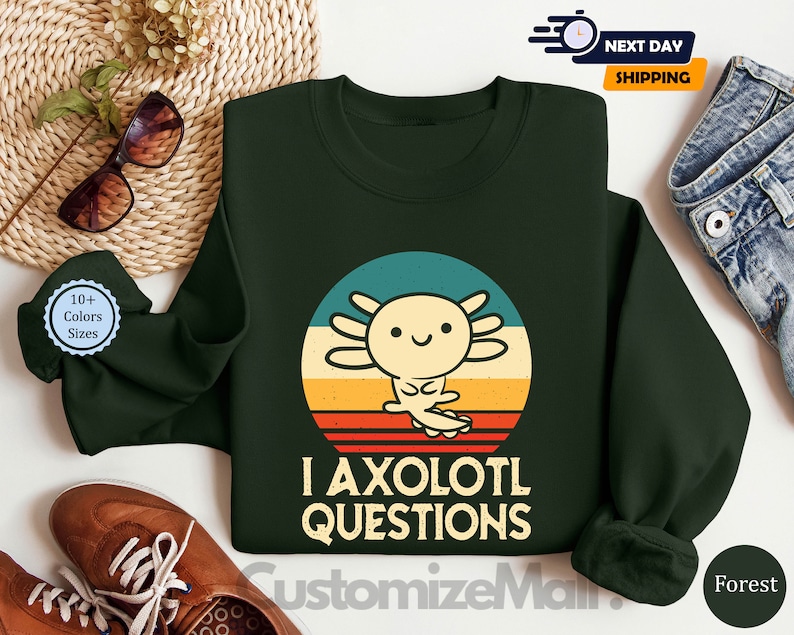 May include: A dark green sweatshirt featuring a retro-style illustration of an axolotl. The axolotl is yellow and has a pink nose. The text "I axolotl questions" is printed below the illustration.