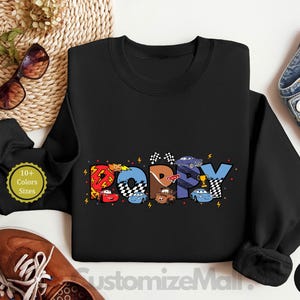 以下が含まれることがあります： カラフルな漫画の車とレースの旗でできた「Happy」という言葉のカラフルなグラフィックが描かれた黒いスウェットシャツ。左下の隅に「10+ Colors Sizes」というテキストが表示されています。