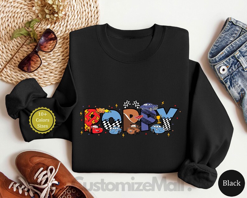 以下が含まれることがあります： カラフルな漫画の車とレースの旗でできた「Happy」という言葉のカラフルなグラフィックが描かれた黒いスウェットシャツ。左下の隅に「10+ Colors Sizes」というテキストが表示されています。