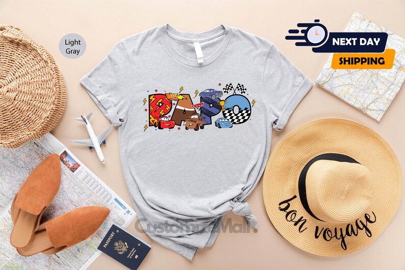 Może przedstawiać: Jasnoszary t-shirt z kolorową grafiką kresk&oacute;wek samochod&oacute;w, kt&oacute;re tworzą słowo "RAPO".