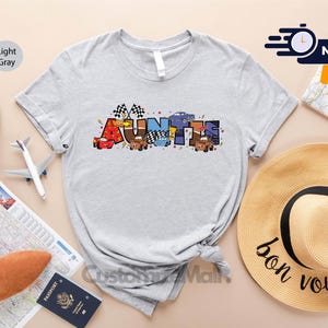 以下が含まれることがあります： 薄いグレーのTシャツに、漫画の車とレースの旗でできた「AUNTIE」という言葉のカラフルなグラフィックが描かれています。