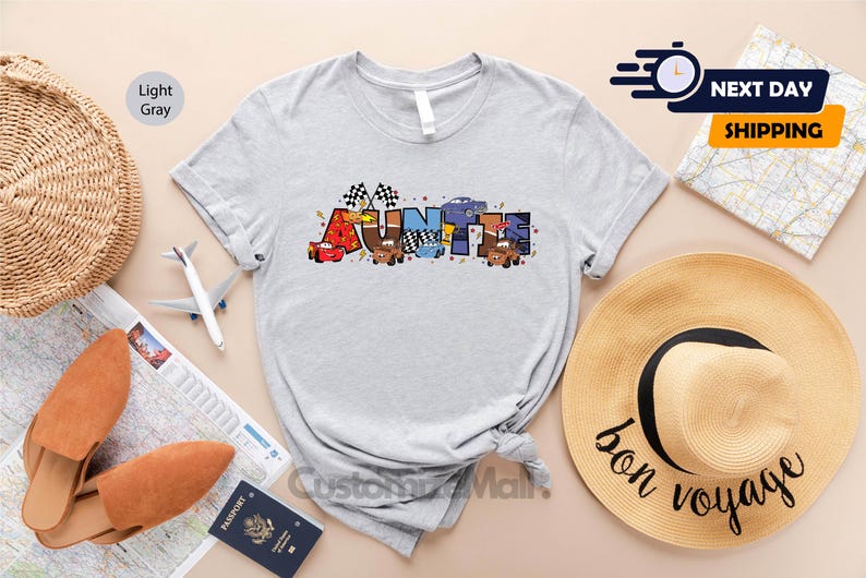 以下が含まれることがあります： 薄いグレーのTシャツに、漫画の車とレースの旗でできた「AUNTIE」という言葉のカラフルなグラフィックが描かれています。