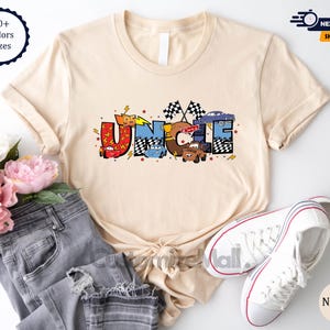 以下が含まれることがあります： ディズニーピクサー映画カーズの漫画の車から作られた「UNCLE」という言葉のグラフィックが描かれたベージュのTシャツ。グラフィックは赤、青、黄、黒です。