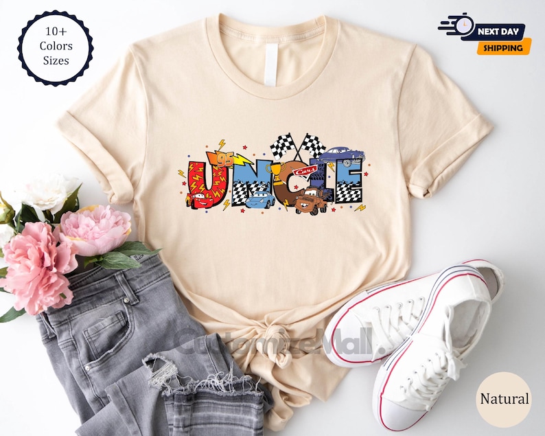 以下が含まれることがあります： ディズニーピクサー映画カーズの漫画の車から作られた「UNCLE」という言葉のグラフィックが描かれたベージュのTシャツ。グラフィックは赤、青、黄、黒です。