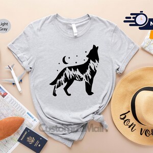 K&ouml;nnte beinhalten: Hellgraues T-Shirt mit einer schwarzen Grafik eines heulenden Wolfs, einem Bergketten-Design, einem Mond und Sternen. Das T-Shirt ist Teil einer Kollektion zum Thema Reisen. Ein Strohhut mit dem Aufdruck "bon voyage" ist ebenfalls zu sehen.