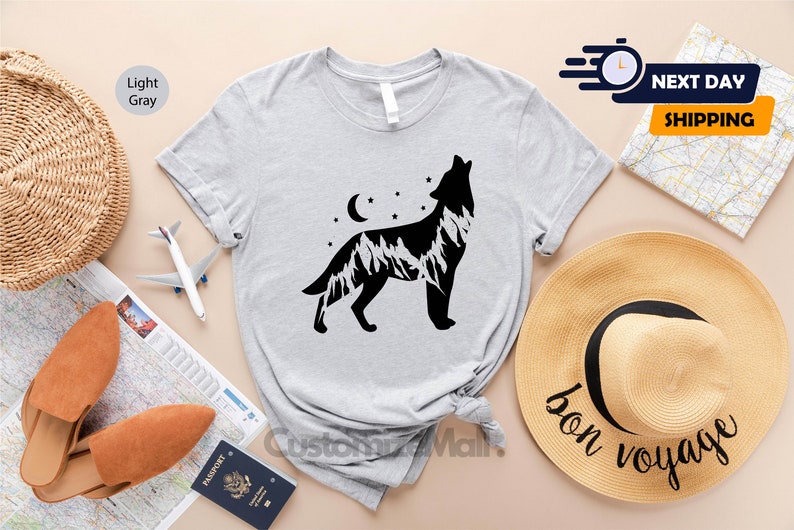 K&ouml;nnte beinhalten: Hellgraues T-Shirt mit einer schwarzen Grafik eines heulenden Wolfs, einem Bergketten-Design, einem Mond und Sternen. Das T-Shirt ist Teil einer Kollektion zum Thema Reisen. Ein Strohhut mit dem Aufdruck "bon voyage" ist ebenfalls zu sehen.
