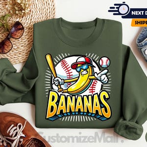 K&ouml;nnte beinhalten: Milit&auml;rgr&uuml;nes Sweatshirt mit Baseball-Motiv. Das Motiv zeigt eine gelbe Banane mit Sonnenbrille und Baseballkappe, die einen Schl&auml;ger h&auml;lt, mit dem Wort "BANANAS" in fetten Buchstaben. Ein Baseball ist ebenfalls im Motiv enthalten.