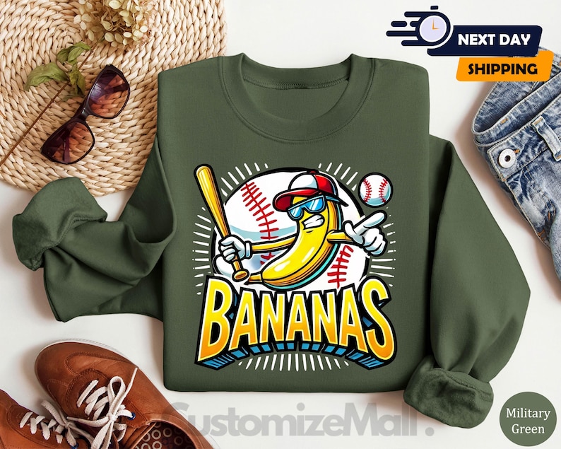 K&ouml;nnte beinhalten: Milit&auml;rgr&uuml;nes Sweatshirt mit Baseball-Motiv. Das Motiv zeigt eine gelbe Banane mit Sonnenbrille und Baseballkappe, die einen Schl&auml;ger h&auml;lt, mit dem Wort "BANANAS" in fetten Buchstaben. Ein Baseball ist ebenfalls im Motiv enthalten.