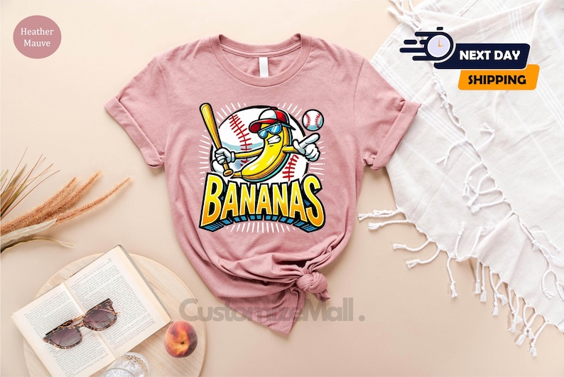 K&ouml;nnte beinhalten: Hellmauvefarbenes T-Shirt mit einer Comic-Banane, die eine Sonnenbrille und eine Baseballkappe tr&auml;gt, einen Schl&auml;ger h&auml;lt und zeigt. Das Wort "BANANAS" ist in einer blauen und gelben 3D-Schriftart gehalten. Ein Baseball und Baseballn&auml;hte sind im Hintergrund.