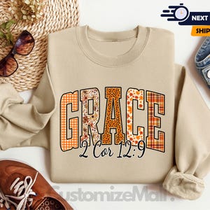 May include: A sand-colored sweatshirt with the word "GRACE" in large, colorful, patterned letters. The patterns include floral, leopard print, and plaid designs. Below the word "GRACE" is the text "2 Cor 12:9".