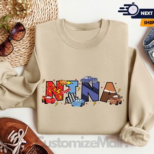 以下が含まれることがあります： カラフルな文字で「NINA」と綴られ、漫画の車とレースの旗が描かれたタン色のスウェットシャツ。柔らかく快適な素材で作られています。