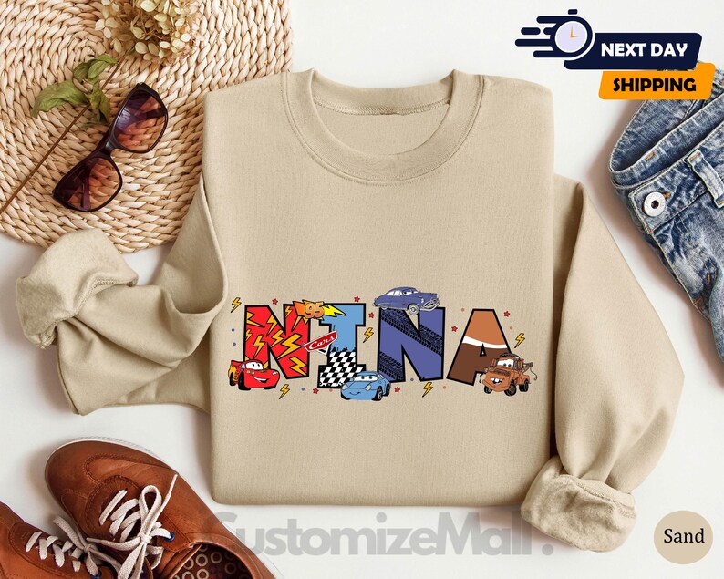 以下が含まれることがあります： カラフルな文字で「NINA」と綴られ、漫画の車とレースの旗が描かれたタン色のスウェットシャツ。柔らかく快適な素材で作られています。