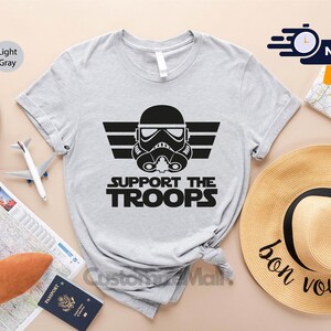 May include: Light gray t-shirt with a black graphic of a Star Wars Stormtrooper helmet with the text "Support the Troops" underneath.