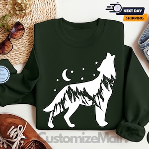 K&ouml;nnte beinhalten: Dunkelgr&uuml;nes Sweatshirt mit einer wei&szlig;en Wolfsilhouette, die Mond und Sterne anheult. Der K&ouml;rper des Wolfes ist mit einem Bergketten-Design gef&uuml;llt. Das Sweatshirt hat einen Rundhalsausschnitt und lange &Auml;rmel. Der Text "CustomizeMain" ist unten sichtbar.