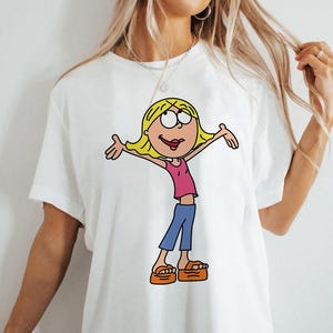 May include: White t-shirt featuring a cartoon illustration of a girl with blonde hair, wearing a pink shirt, blue trousers, and orange sandals.