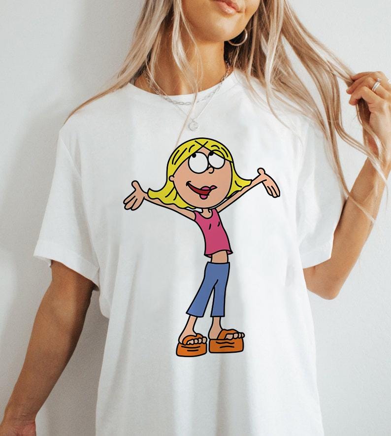 May include: White t-shirt featuring a cartoon illustration of a girl with blonde hair, wearing a pink shirt, blue trousers, and orange sandals.