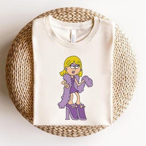 May include: A white t-shirt featuring a cartoon illustration of a girl with blonde hair wearing a purple dress and purple feather boa. The girl is wearing purple high heels and has a surprised expression on her face.