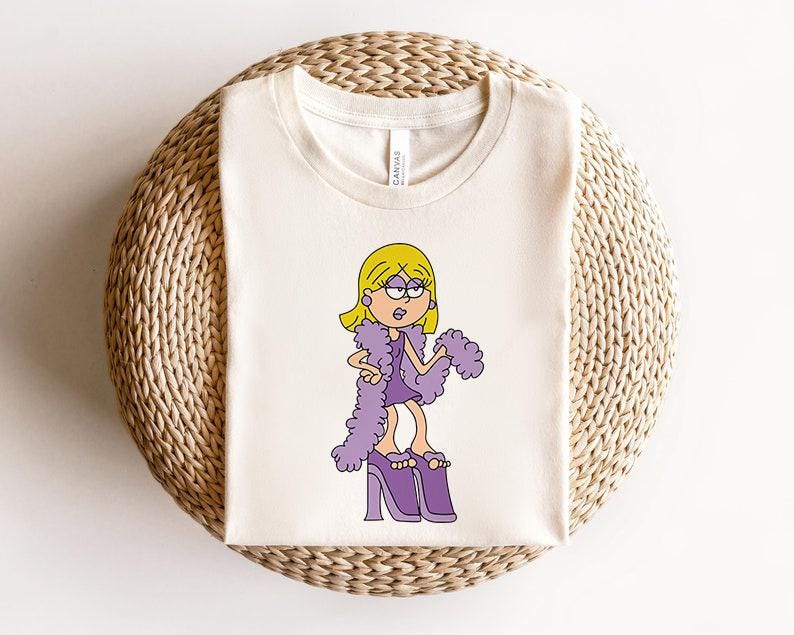 May include: A white t-shirt featuring a cartoon illustration of a girl with blonde hair wearing a purple dress and purple feather boa. The girl is wearing purple high heels and has a surprised expression on her face.