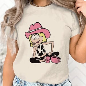 May include: A white t-shirt featuring a cartoon illustration of a girl wearing a pink cowboy hat and a cow print top. The girl is sitting with her legs crossed and has a thoughtful expression on her face.
