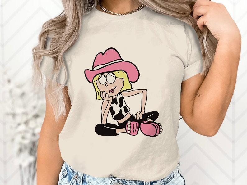 May include: A white t-shirt featuring a cartoon illustration of a girl wearing a pink cowboy hat and a cow print top. The girl is sitting with her legs crossed and has a thoughtful expression on her face.