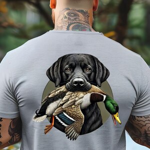 Pu&ograve; includere: T-shirt grigio chiaro con un disegno di un Labrador Retriever nero che tiene un'anatra in bocca. L'anatra ha piume marroni, bianche e blu, una testa verde e un becco giallo. Il cane &egrave; su un cerchio verde.