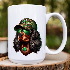 May include: White ceramic mug with a graphic of a black and brown dog wearing a camouflage hat with the text "Lucky Dog" and green sunglasses. The dog is wearing a green shamrock necklace.