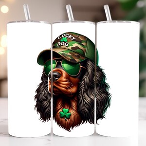 May include: A white tumbler with a graphic of a brown and black dog wearing a green camouflage hat with the words "Lucky Dog" on it. The dog is also wearing green sunglasses and a shamrock.