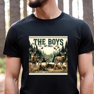 K&ouml;nnte beinhalten: Ein schwarzes T-Shirt mit einer Grafik im Vintage-Stil von einer Gruppe Hirsche in einem Wald. Die Grafik lautet "The Boys of Fall".