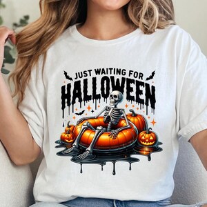May include: White t-shirt with a black graphic of a skeleton sitting in a pumpkin-shaped float. The text "Just Waiting for Halloween" is above the skeleton.
