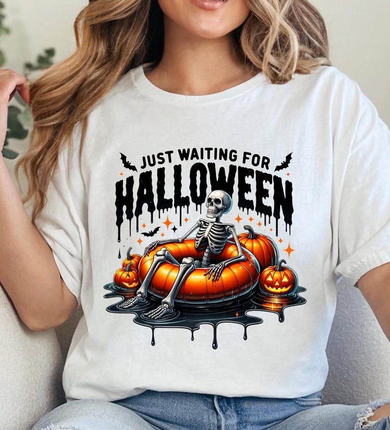 May include: White t-shirt with a black graphic of a skeleton sitting in a pumpkin-shaped float. The text "Just Waiting for Halloween" is above the skeleton.