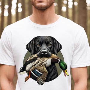 Pu&ograve; includere: T-shirt bianca con un disegno di un Labrador Retriever nero che tiene un'anatra in bocca. L'anatra ha piume marroni, bianche, verdi e gialle. Il cane &egrave; su uno sfondo circolare verde.