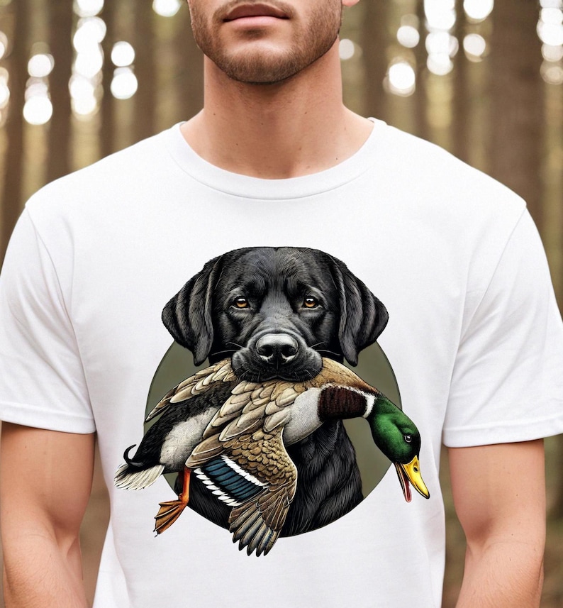 Pu&ograve; includere: T-shirt bianca con un disegno di un Labrador Retriever nero che tiene un'anatra in bocca. L'anatra ha piume marroni, bianche, verdi e gialle. Il cane &egrave; su uno sfondo circolare verde.